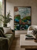 A large framed abstract landscape painting hangs on a textured beige wall in a styled living room with a green velvet sofa, a light cream armchair, and a low stone coffee table.